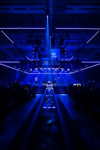 A dimly lit stage surrounded by a large audience, with blue lighting creating an atmospheric ambiance. The stage features several performers and screens displaying images, with a central light beam illuminating a portion of the stage.