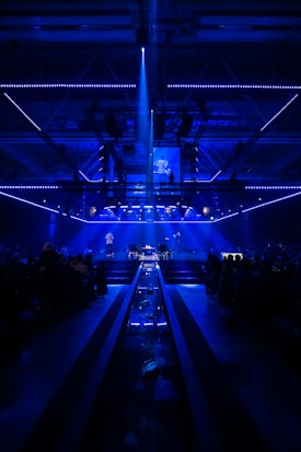 A dimly lit stage surrounded by a large audience, with blue lighting creating an atmospheric ambiance. The stage features several performers and screens displaying images, with a central light beam illuminating a portion of the stage.