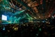 A large conference or event is taking place in a spacious hall with an audience seated in rows facing a well-lit stage. The stage has a screen displaying text and a presenter standing beside it. The ceiling is equipped with numerous lights and trusses, creating a dynamic atmosphere with green and blue lighting effects.