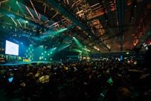 A large conference or event is taking place in a spacious hall with an audience seated in rows facing a well-lit stage. The stage has a screen displaying text and a presenter standing beside it. The ceiling is equipped with numerous lights and trusses, creating a dynamic atmosphere with green and blue lighting effects.