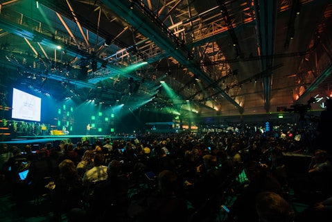 A large conference or event is taking place in a spacious hall with an audience seated in rows facing a well-lit stage. The stage has a screen displaying text and a presenter standing beside it. The ceiling is equipped with numerous lights and trusses, creating a dynamic atmosphere with green and blue lighting effects.