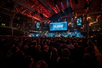A large audience is gathered in a dimly lit auditorium with vibrant red and blue lighting. A prominent stage features a big screen displaying the word 'SLUSH'. Multiple smaller screens surround the main stage showing various images or presentations. The room has an industrial, tech-friendly vibe with many attendees seated, facing the stage.