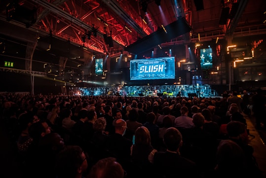 A large audience is gathered in a dimly lit auditorium with vibrant red and blue lighting. A prominent stage features a big screen displaying the word 'SLUSH'. Multiple smaller screens surround the main stage showing various images or presentations. The room has an industrial, tech-friendly vibe with many attendees seated, facing the stage.