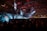 A large indoor event with a stage featuring bright screens and spotlights, attended by a substantial crowd. The venue has a high ceiling with industrial beams and red lighting accents. The atmosphere resembles a concert or a conference with a focus on visual effects.