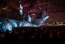 A large indoor event with a stage featuring bright screens and spotlights, attended by a substantial crowd. The venue has a high ceiling with industrial beams and red lighting accents. The atmosphere resembles a concert or a conference with a focus on visual effects.