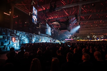 A large indoor event with a stage featuring bright screens and spotlights, attended by a substantial crowd. The venue has a high ceiling with industrial beams and red lighting accents. The atmosphere resembles a concert or a conference with a focus on visual effects.