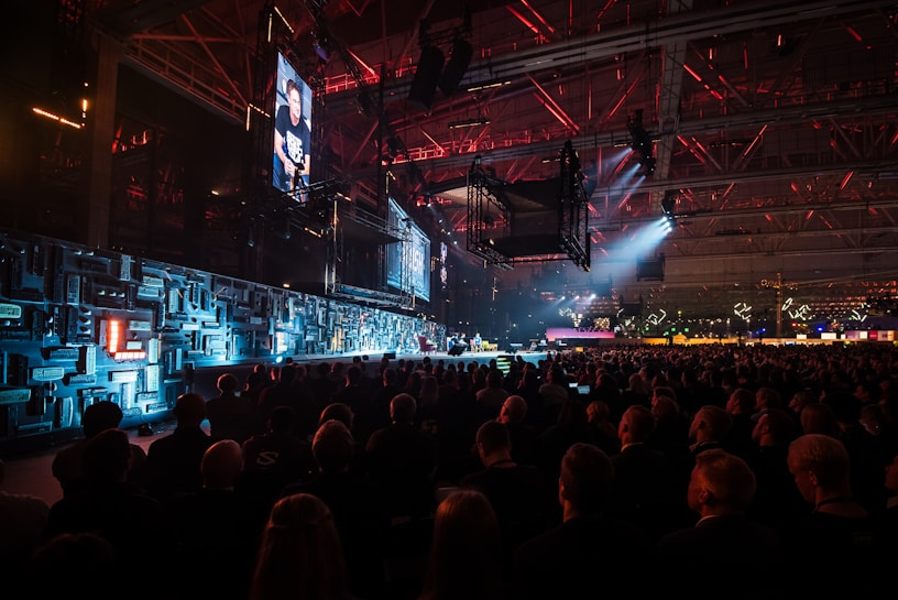 A large indoor event with a stage featuring bright screens and spotlights, attended by a substantial crowd. The venue has a high ceiling with industrial beams and red lighting accents. The atmosphere resembles a concert or a conference with a focus on visual effects.
