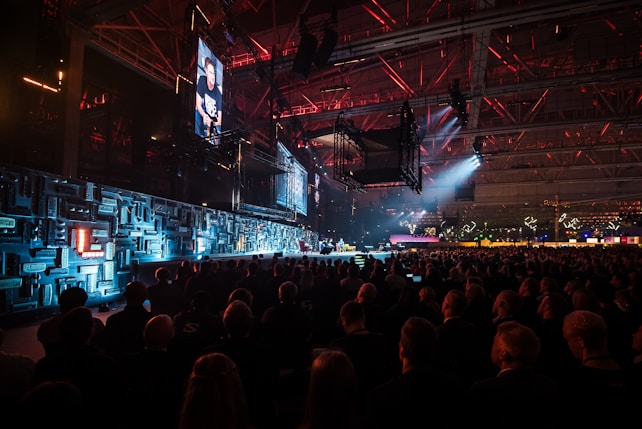 A large indoor event with a stage featuring bright screens and spotlights, attended by a substantial crowd. The venue has a high ceiling with industrial beams and red lighting accents. The atmosphere resembles a concert or a conference with a focus on visual effects.