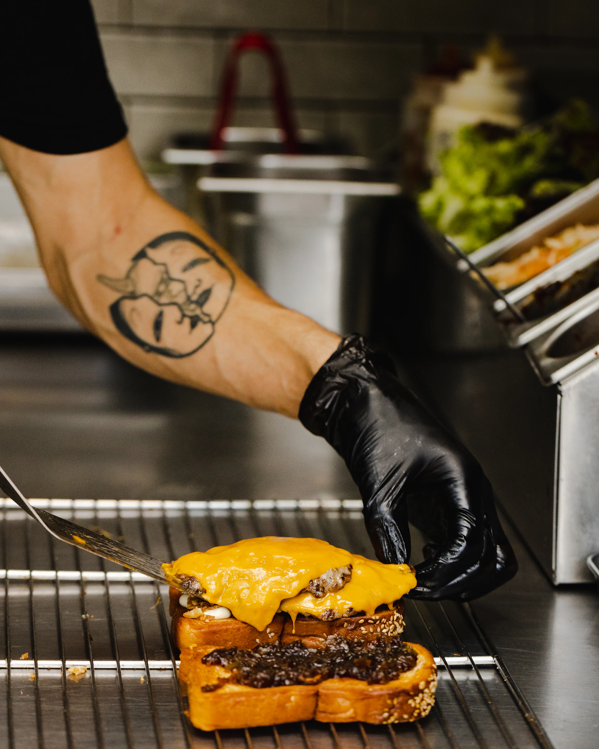 A person wearing a black glove prepares a sandwich in a kitchen setting. The sandwich features two slices of bread, melted cheddar cheese, and a beef patty with what appears to be caramelized onions. The forearm of the person shows a distinct tattoo, and various kitchen items like a tray of lettuce are visible in the background.