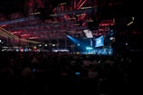 A large crowd is seated inside an industrial-looking venue with high ceilings. The space is dimly lit with overhead red and blue lights, creating a captivating atmosphere. A prominent screen displaying the word 'SLUSH' is visible, indicating a technology or startup conference setting.