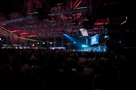 A large crowd is seated inside an industrial-looking venue with high ceilings. The space is dimly lit with overhead red and blue lights, creating a captivating atmosphere. A prominent screen displaying the word 'SLUSH' is visible, indicating a technology or startup conference setting.