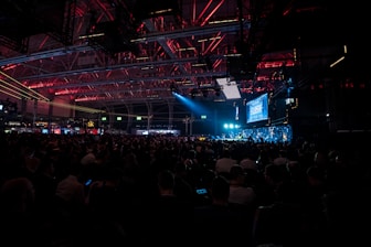 A large crowd is seated inside an industrial-looking venue with high ceilings. The space is dimly lit with overhead red and blue lights, creating a captivating atmosphere. A prominent screen displaying the word 'SLUSH' is visible, indicating a technology or startup conference setting.