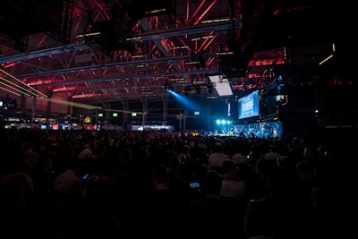 A large crowd is seated inside an industrial-looking venue with high ceilings. The space is dimly lit with overhead red and blue lights, creating a captivating atmosphere. A prominent screen displaying the word 'SLUSH' is visible, indicating a technology or startup conference setting.