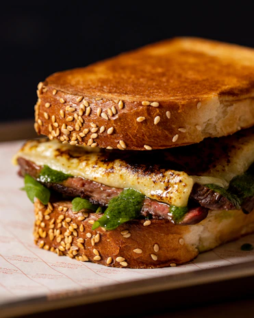 A gourmet sandwich made with thick slices of toasted bread covered with sesame seeds. Inside, there is a layer of grilled meat complemented by a melted cheese topping and a vibrant green sauce, possibly pesto or chimichurri.