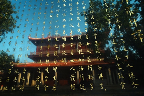 Golden script with intricate calligraphy overlays a traditional Asian architectural structure featuring red roofs and white columns. Trees frame the composition, and the reflection creates a layered visual effect.