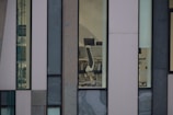 Several large windows reveal a sparse office interior with modern white office chairs and black monitors against a backdrop of a minimalist, multi-textured building facade.