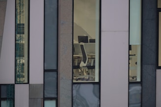 Several large windows reveal a sparse office interior with modern white office chairs and black monitors against a backdrop of a minimalist, multi-textured building facade.