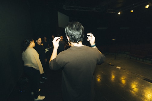 A group of people stand on a dimly lit stage, possibly rehearsing. The focus is on a person holding a microphone, gesturing with their hands, while others are scattered around, some appearing to hold musical instruments.