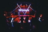 A dimly lit stage with vibrant red and blue lighting features a performer in front of an audience. The backdrop displays bold white text that reads 'ES PARA TI'. Spotlights project beams of light onto the stage, creating a lively atmosphere, while silhouettes of people can be seen enjoying the performance.