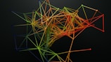A complex, abstract structure composed of interconnected lines and nodes, forming a colorful network on a dark background. The lines are primarily shades of green, yellow, orange, and red, creating a gradient effect across the structure.