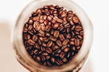 Coffee beans are seen inside a glass jar, with a close-up perspective highlighting their rich, brown tones and glossy textures.