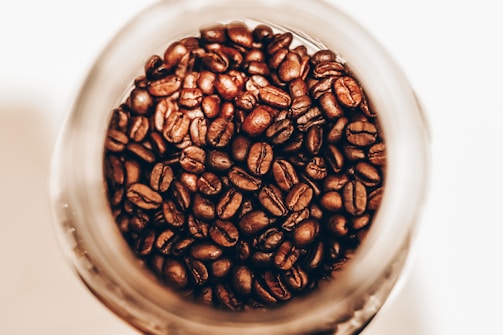 Coffee beans are seen inside a glass jar, with a close-up perspective highlighting their rich, brown tones and glossy textures.