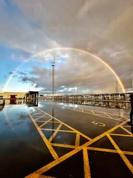 A large, vivid rainbow arches across a cloudy sky over an industrial area, with reflections visible on the wet pavement. Yellow road markings are prominent in the foreground, while a series of poles and buildings are seen in the background.