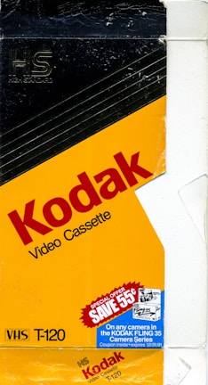 A vintage Kodak video cassette packaging with a predominantly yellow and black design. The prominent red text spells 'Kodak Video Cassette', while the bottom left corner displays 'VHS T-120'. There is a blue promotional label offering a discount on Kodak Fling 35 Camera Series, valid until 1991.