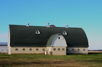 A large, light-colored barn with a curved, dark-green roof sits in a grassy field on a clear day. The barn features multiple small windows along the base and three cupolas on the roof. There are a few small structures and objects near the barn's side, including propane tanks.