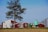 A rural scene featuring a farm with a red and white barn, a silo, and a green tractor parked nearby. In the foreground, there is a large tree and a light green water tank. The ground shows a field with harvested crops, and the sky is clear with a few clouds.
