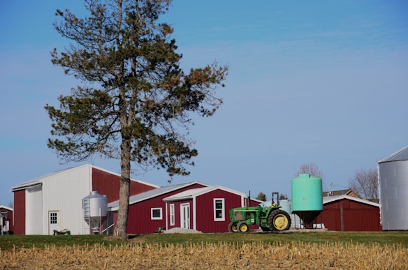 A rural scene featuring a farm with a red and white barn, a silo, and a green tractor parked nearby. In the foreground, there is a large tree and a light green water tank. The ground shows a field with harvested crops, and the sky is clear with a few clouds.
