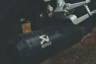 A close-up of a motorcycle exhaust system featuring the brand Akrapovič. The metallic parts, including the footrest and surrounding mechanical components, are visible, displaying intricate details of the engineering. The exhaust appears slightly weathered, with dirt or grime marks visible on the surface.