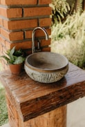 A rustic outdoor sink setup featuring a round, stone basin and a simple metal faucet installed on a sturdy wooden counter. The setup is adjacent to a brick column, and a small potted plant adds a touch of greenery.