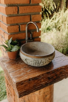 A rustic outdoor sink setup featuring a round, stone basin and a simple metal faucet installed on a sturdy wooden counter. The setup is adjacent to a brick column, and a small potted plant adds a touch of greenery.