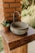 A rustic outdoor sink setup featuring a round, stone basin and a simple metal faucet installed on a sturdy wooden counter. The setup is adjacent to a brick column, and a small potted plant adds a touch of greenery.