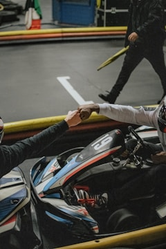 Two individuals wearing helmets engage in a fist bump while seated in go-karts on an indoor track. The image captures a moment of camaraderie in a competitive setting, with a focus on the go-kart adorned with the number 9 and sporting a sleek design with racing stripes. A third person stands nearby holding an item, partially visible in the background.