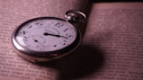 A vintage pocket watch with Roman numerals rests on the open pages of an old book. The book's pages contain text in a serif font, and the watch's metal casing is slightly tarnished, suggesting age. The timepiece is positioned diagonally, with the winding crown facing up, casting a soft shadow on the paper.