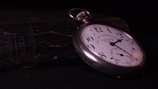 A vintage pocket watch lies atop a weathered, textured book. The watch has a white face with black numerals and a silver casing. The watch hands point to approximately 10:10. The background is dark, highlighting the watch and creating a sense of timelessness.