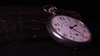 A vintage pocket watch lies atop a weathered, textured book. The watch has a white face with black numerals and a silver casing. The watch hands point to approximately 10:10. The background is dark, highlighting the watch and creating a sense of timelessness.