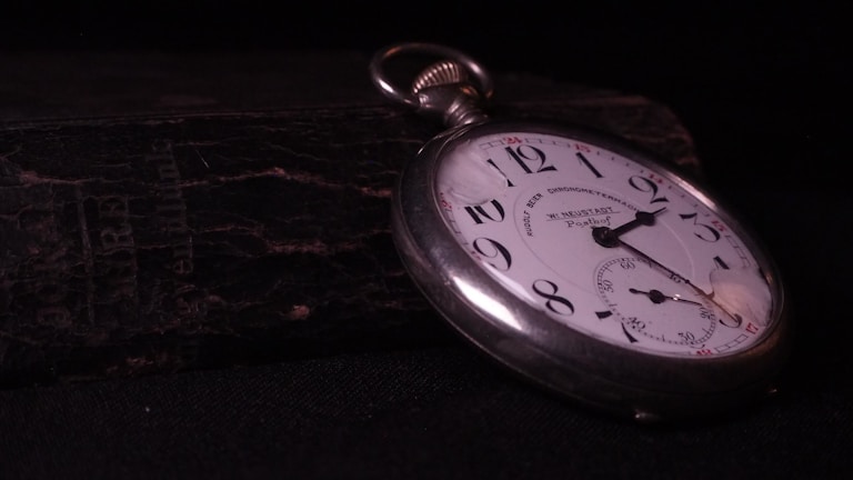 A vintage pocket watch lies atop a weathered, textured book. The watch has a white face with black numerals and a silver casing. The watch hands point to approximately 10:10. The background is dark, highlighting the watch and creating a sense of timelessness.