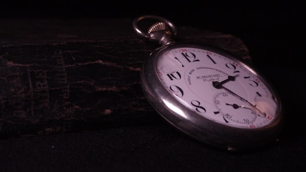 A vintage pocket watch lies atop a weathered, textured book. The watch has a white face with black numerals and a silver casing. The watch hands point to approximately 10:10. The background is dark, highlighting the watch and creating a sense of timelessness.