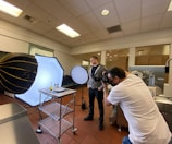 A photographer is taking pictures of a food setup in a studio or kitchen environment. The scene includes professional lighting equipment, such as softboxes and reflectors. Two individuals are present, with one focusing on capturing the image while the other stands nearby.