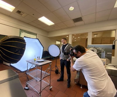A photographer is taking pictures of a food setup in a studio or kitchen environment. The scene includes professional lighting equipment, such as softboxes and reflectors. Two individuals are present, with one focusing on capturing the image while the other stands nearby.
