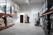 A spacious warehouse aisle with shelves stacked high with pallets wrapped in clear plastic. Boxes of various sizes are stored on orange metal racks, and a man is standing at the back wall near a stack of boxes. A small forklift labeled Toyota is positioned nearby.