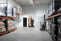 A spacious warehouse aisle with shelves stacked high with pallets wrapped in clear plastic. Boxes of various sizes are stored on orange metal racks, and a man is standing at the back wall near a stack of boxes. A small forklift labeled Toyota is positioned nearby.