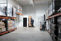 A spacious warehouse aisle with shelves stacked high with pallets wrapped in clear plastic. Boxes of various sizes are stored on orange metal racks, and a man is standing at the back wall near a stack of boxes. A small forklift labeled Toyota is positioned nearby.