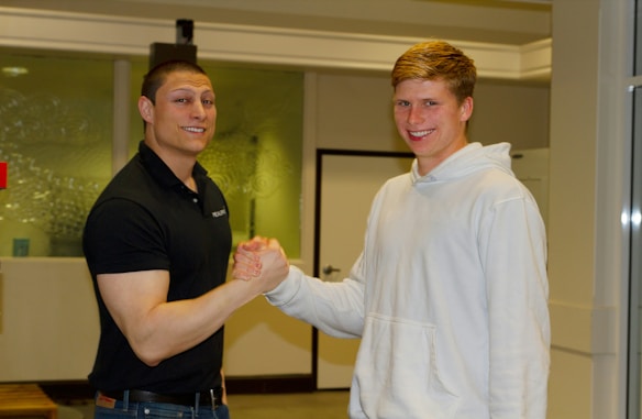 Two people are indoors, shaking hands with a friendly expression. One is wearing a black polo shirt and the other a white hoodie. They stand in front of a glass wall with a door visible in the background.