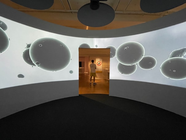 A person stands in a hallway illuminated by a backlit projection of abstract, circular shapes resembling bubbles or orbs. The walls around them are curved and display the same projection, creating an immersive environment.