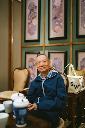 An elderly person wearing a navy blue jacket sits in a room decorated with elegant wall panels featuring floral and bird artwork. To the left, there are several disposable coffee cups on a wooden table, and beside the person, a shopping bag with the word 'SWE' is visible.
