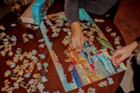 Two people are assembling a colorful jigsaw puzzle on a wooden table. The partially completed puzzle features a scenic image with buildings and a road. Several loose puzzle pieces are scattered around the table, and a puzzle box is placed nearby.